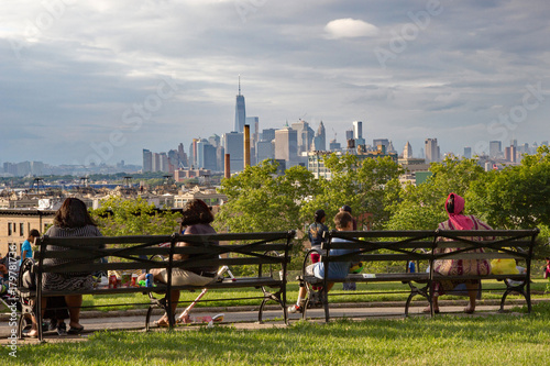 New York's diversity represented by people sat on a bench in Sunset Park, Brooklyn, admiring the view of Lower Manhattan on a sunny summer's day