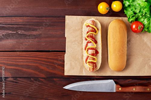 Tasty Hot Dog with sausage, tomatoes, salad, and knife flat lay on wooden background. Top view.