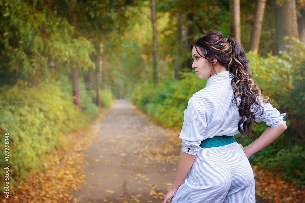 Brunette hairstyle with Greek braid in white stylish dress at the background of the road in the Park from behind Brunette hairstyle with Greek braid in white stylish dress at the background of the road in the Park from behind