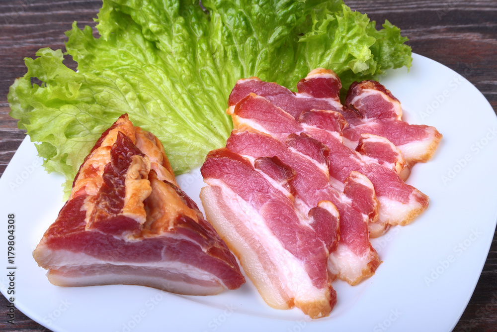 Fresh homemade smoked bacon with leaves lettuce on white plate. Selective focus.