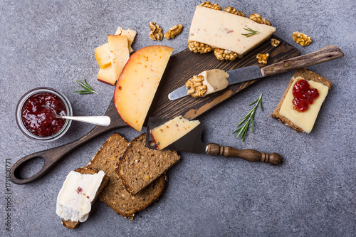 Various types of cheese with wholegrain bread, strawberry jam and walnuts on wooden board on gray stone background. Top view, flat lay, copy space.