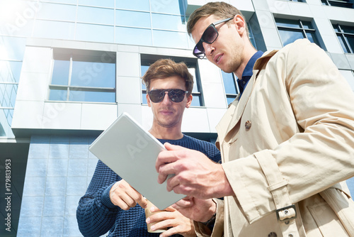 Low angle portrait of two young entrepreneurs using digital tablet outdoors and discussing work standing against glass front of modern office building, copy space