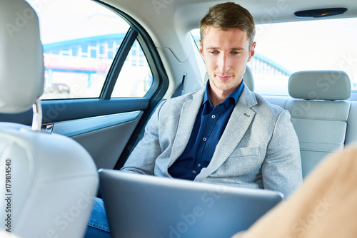 Portrait of successful young businessman using laptop for work sitting in back seat of  luxury car, copy space © Seventyfour