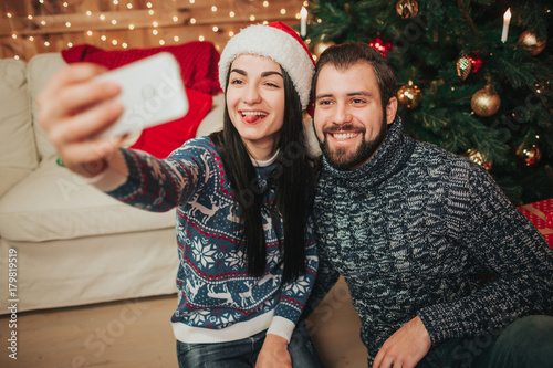 Merry Christmas and Happy New Year!. Young couple celebrating holiday at home. Man and woman do selfie on smartphone