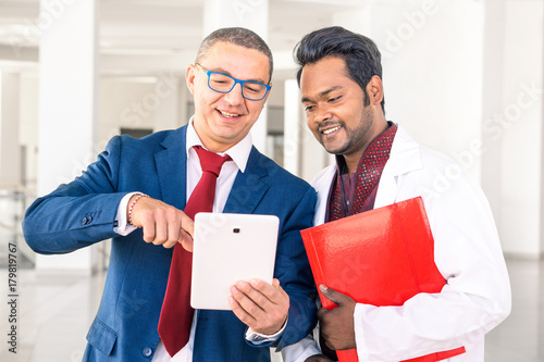 Young Indian doctor with businessman talking in hospital hallway - Salesman holding pad showing health care equipment at asian medical director - Concept of sales, business, deals with multiracial men