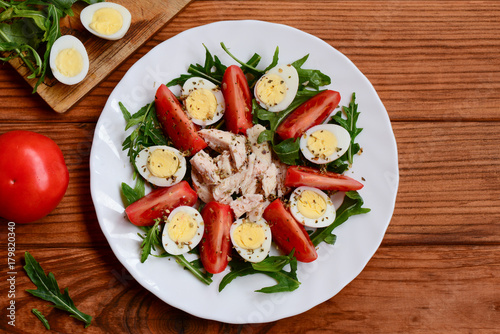 Easy vegetable chicken salad idea for lunch or dinner. Home salad with fresh tomatoes, arugula, quail eggs, chicken breast and spices on a white plate and a wooden table. Closeup. Top view