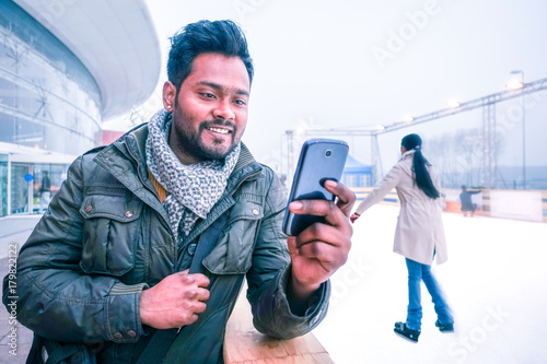 Happy indian man looking mobile phone outdoors at winter with ice skating scene background - Cheerful bangladeshi young guy using cellphone smiling looking telephone screen outside on grey light day photo