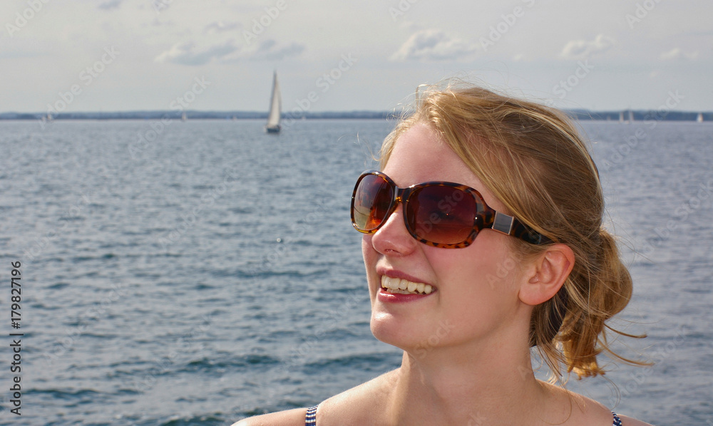Portrait of a beautiful young blond woman wearing sunglasses with the sea and a distant sailboat in the background Portrait of a beautiful young blond woman wearing sunglasses with the sea and a distant sailboat in the background