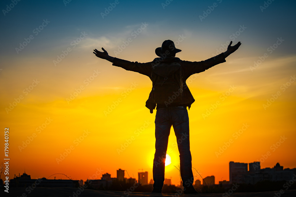 Traveler silhouette with a backpack and cowboy hat stand on hill top. City in the background. Sun sets over horizon. Man looks ahead, raise his arms up. Travel, holidays, advanture, success concept.