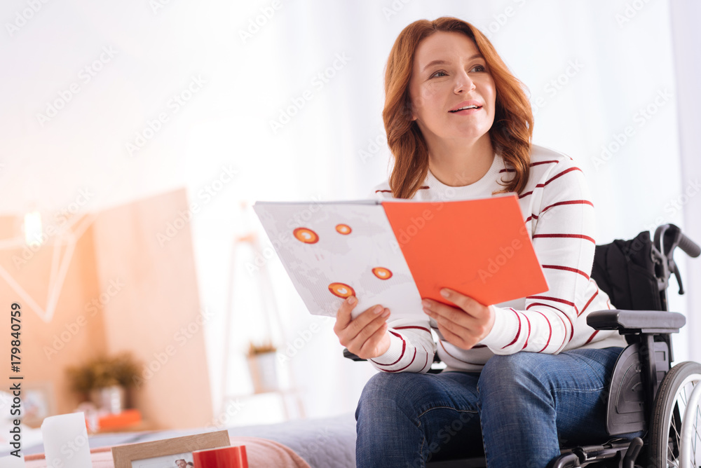 I like reading. Good-looking alert blond disabled woman of middle age smiling and holding a notebook while sitting in the wheelchair