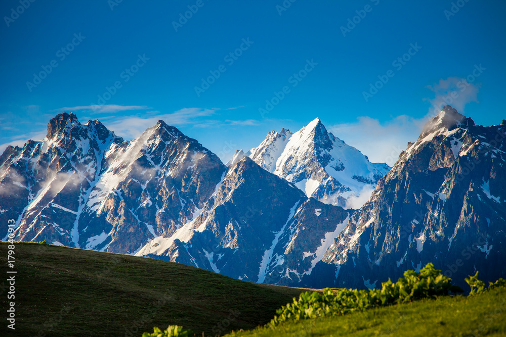 Panoramic summer landscape with green hill and mountain snowy peaks against clear blue sky. Svanetia region, Georgia. Main Caucasian ridge. Travel background. Holiday, hiking, sport, recreation.
