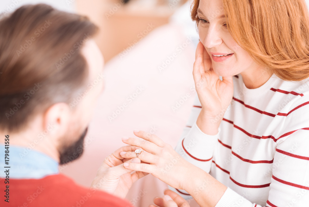 Loving each other. Beautiful cheerful blond woman of middle age smiling and looking at her future husband while he putting the wedding ring on her finger Loving each other. Beautiful cheerful blond woman of middle age smiling and looking at her future husband while he putting the wedding ring on her finger