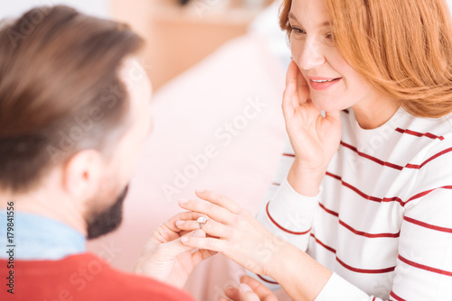 Loving each other. Beautiful cheerful blond woman of middle age smiling and looking at her future husband while he putting the wedding ring on her finger