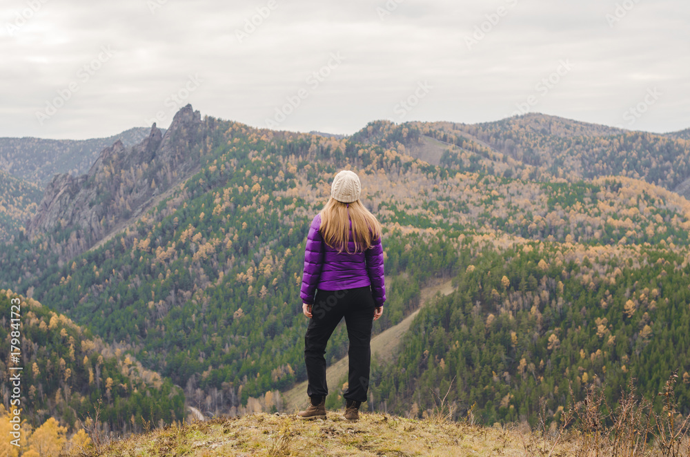 A girl in a lilac jacket standing on a mountain, a view of the mountains and an autumn forest by a cloudy day A girl in a lilac jacket standing on a mountain, a view of the mountains and an autumn forest by a cloudy day
