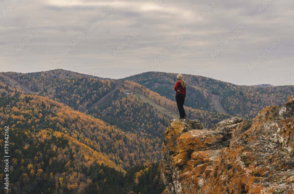 A girl in a red jacket looks out into the distance on a mountain, a view of the mountains and an autumnal forest by an overcast day. Free space for text
