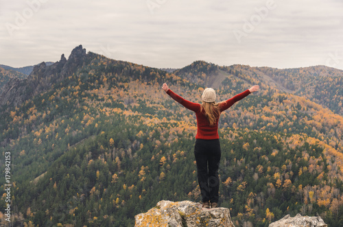 A girl in a red jacket stretching her arms on a mountain, a view of the mountains and an autumn forest by a cloudy day, free space for text
