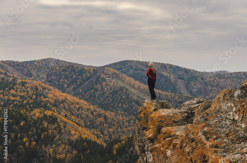 A girl in a red jacket looks out into the distance on a mountain, a view of the mountains and an autumnal forest by an overcast day. Free space for text © Maksim