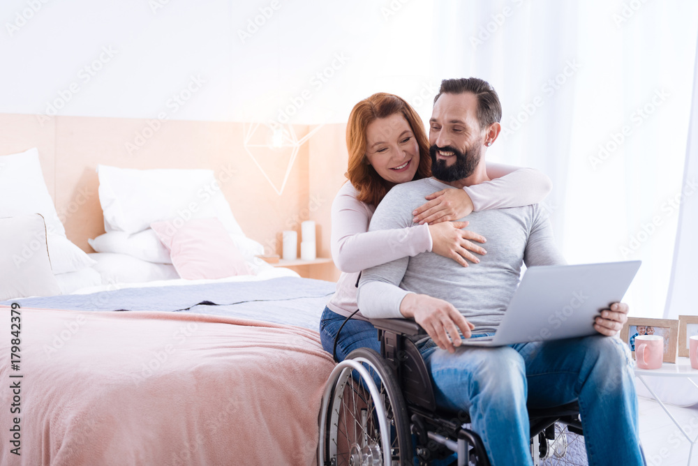 We love cuddling. Blond woman of middle age and a bearded disabled man smiling while the woman hugging him and the man sitting in a wheelchair with a laptop