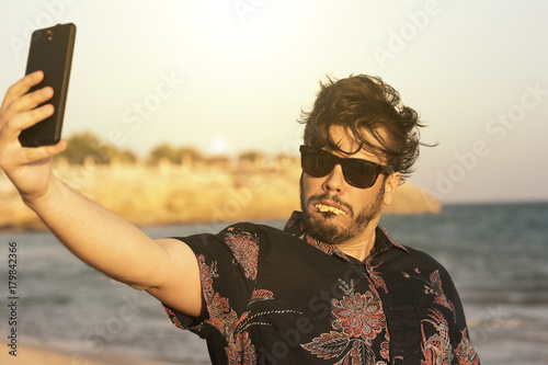 O homem feio usa camisa azul florida e oculos no dia ensolarado e quente na praia. Homem engraçado fazendo selfie com o seu telefone celular smart. photo