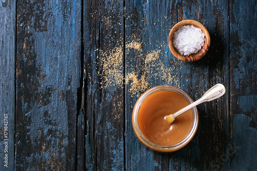 Glass jar of homemade salted caramel sauce with spoon, brown sugar and bowl of salt. Over old dark blue wooden background. Top view with space photo