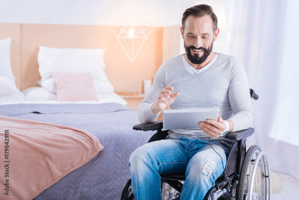 Playing computer games. Content cute bearded disabled man smiling and holding a tablet while sitting in a wheelchair