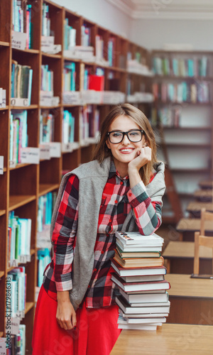 Portrait of a smiling casual student girl wears glasses holding stack of books in evening library, education concept