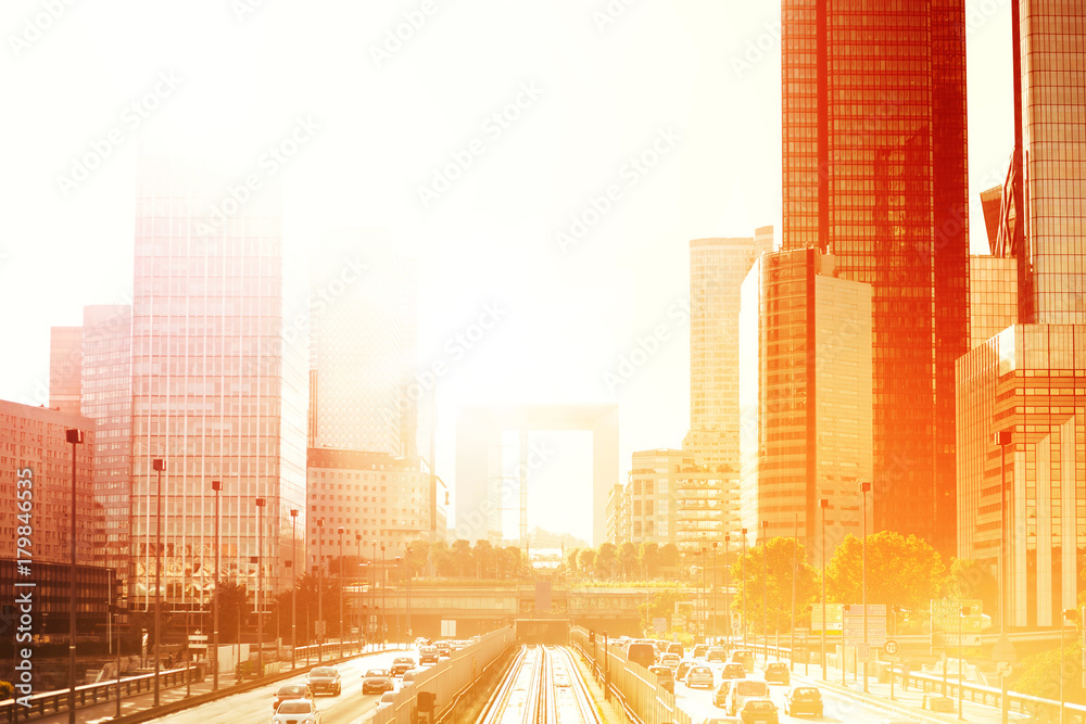 Obraz premium Modern buildings and dynamic street traffic in Paris business district La Defense. Glass facade skyscrapers on a bright sunny day with sunbeams in the sky. Economics, finances and city traffic concept