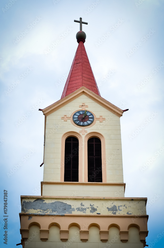 front view of yellow cathedral with cross, clock with Roman numbers and red roof front view of yellow cathedral with cross, clock with Roman numbers and red roof