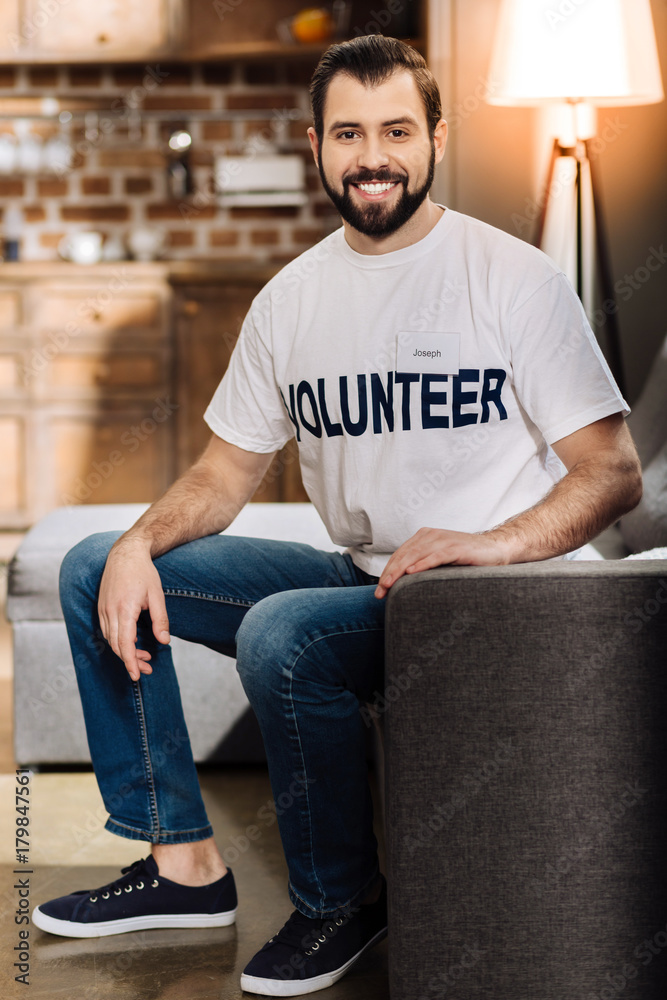 Happy volunteer. Cheerful glad young man being happy while sitting on the sofa and being ready to help people Happy volunteer. Cheerful glad young man being happy while sitting on the sofa and being ready to help people