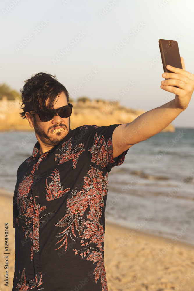 O homem feio usa camisa azul florida e oculos no dia ensolarado e quente na  praia. Homem engraçado fazendo selfie com o seu telefone celular smart.  Stock Photo