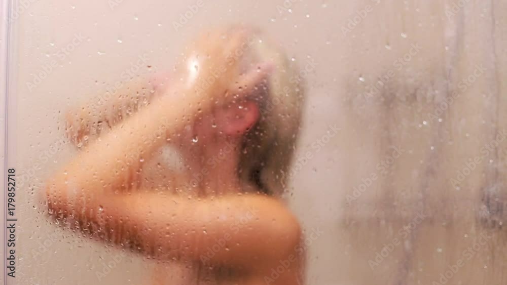 A woman washes her hair in a hot shower behind a glass wall A woman washes her hair in a hot shower behind a glass wall