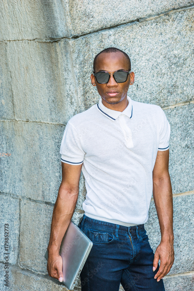 African American College Student studying in New York. Wearing white Polo  shirt, blue jeans, carrying laptop computer, wearing sunglasses, black man  standing by rocky wall on campus, looking forward.. Stock Photo