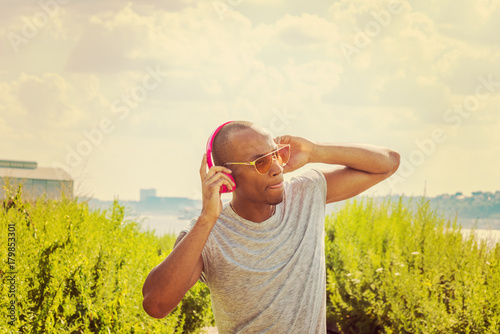 African American Man listening music in New York, wearing sunglasses, raising arms, holding pink wireless headphones, standing by grasses at park under sun, tilting body, biting tongue, looking away.
