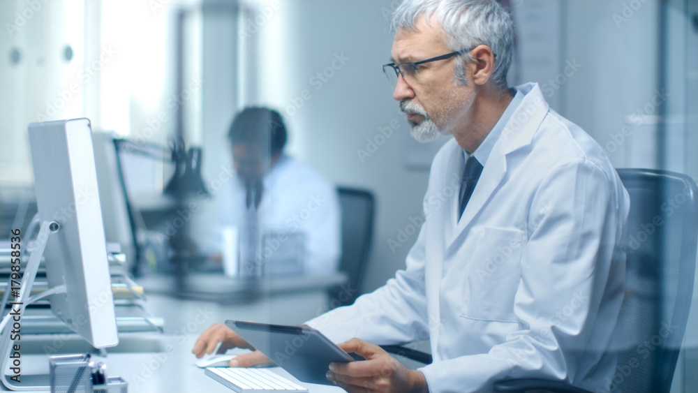 Senior Medical Practitioner Sitting Before Personal Computer Uses Tablet Computer with His One Hand and Operates Keyboard with Other. His Assistant Working in Background. Senior Medical Practitioner Sitting Before Personal Computer Uses Tablet Computer with His One Hand and Operates Keyboard with Other. His Assistant Working in Background.