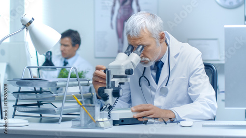 Senior Male Doctor Investigates Health Related Matters Through Microscope. His Assistant Works Nearby at Desktop Computer.