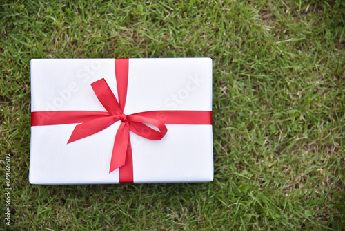 White gift box with red ribbon on green grass background.