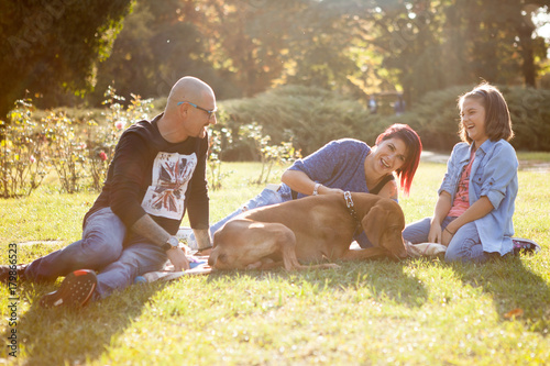 Beautiful laughing family of three playing in the park with their dog. Animal lovers. Mother, father, daughter and their dog
