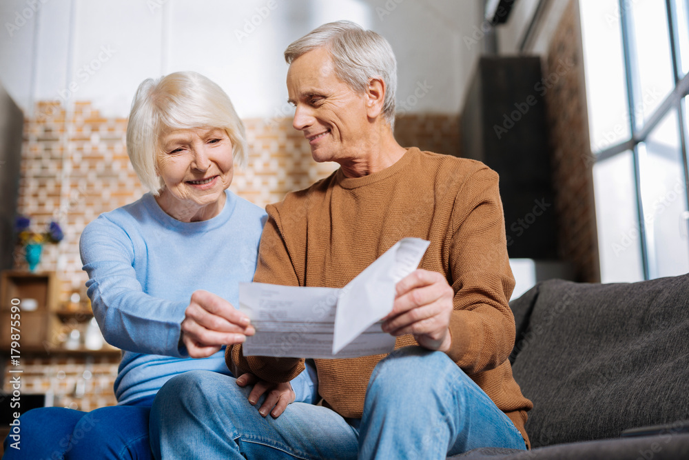 Pleasant notification. Joyful nice elderly woman looking at the piece of paper and smiling while seeing good news Pleasant notification. Joyful nice elderly woman looking at the piece of paper and smiling while seeing good news