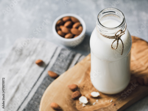 The almond milk in the glass bottle with almond nuts in the white bowl on the wooden decorative rustic cutting board.
