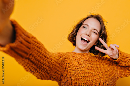 Indoor portrait of blissful girl in knitted clothes making selfie and laughing on bright background. Enchanting young lady posing with peace sign in studio and smiling. photo