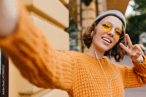 Attractive young woman in sport hat making selfie with peace sign during walk outdoor in cold day. Portrait of cheerful girl in bright knitted sweater having fun on the street.