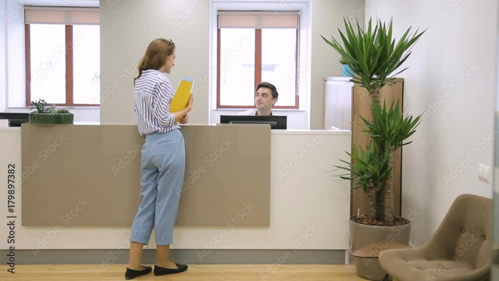 Woman at reception passes documents to man who sits talking on phone. Female employee passes folder with papers to male secretary, who is sitting at counter.
