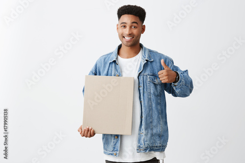 Copy space. Close up of cheerful young black-skinned male student with afro hairstyle in stylish casual outfit holding carton in hand, showing thumb up with happy and satisfied face expression