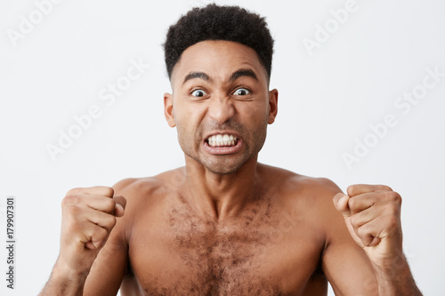 Close up of funny attractive dark-skinned african male with curly hair looking TV with mad expression, cheering for his favorite baseball team, screaming and gesticulating with hands.