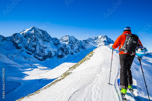 Mountaineer backcountry ski walk along a snowy ridge with skis in the backpack. In background blue sky and shiny sun and Ortler in South Tirol, Italy.