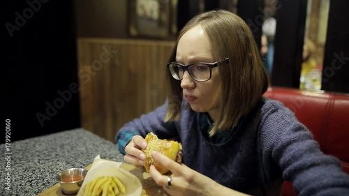 Young girl is eating untasty burger and French fries in the cafe. A woman in glasses is disappointed with the taste of stale cutlets in a hamburger and feel bad. Substandard food products, fast food. photo