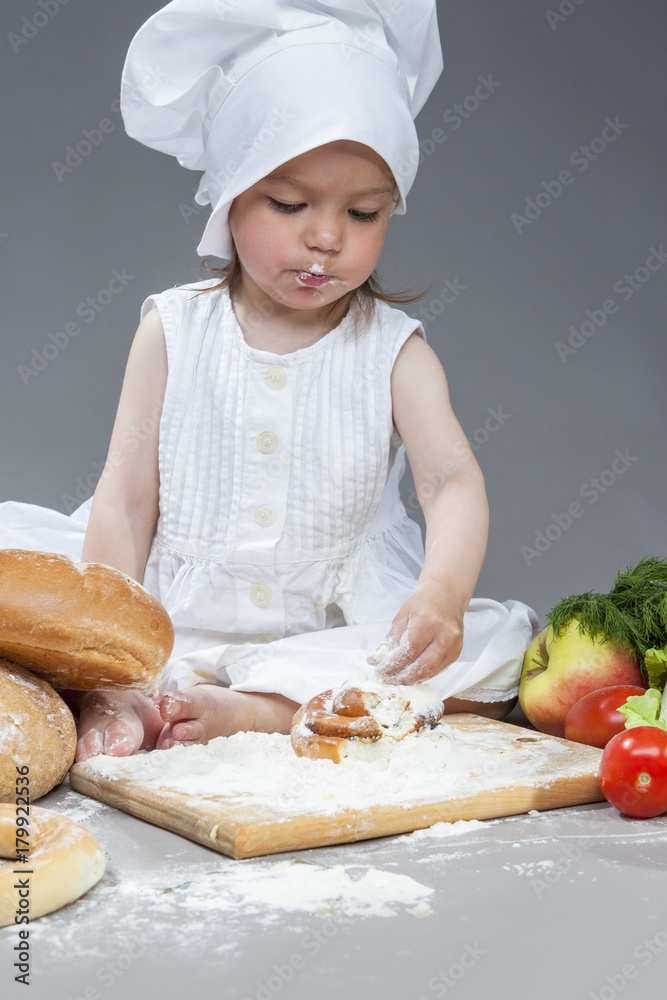 Cooking Ideas and Concepts. Portrait of Little Caucasian Girl in Cook Hat and Lips in Flour Sitting in Front of Fresh Buns. Sieving Powder on Pastry. Against Gray. Cooking Ideas and Concepts. Portrait of Little Caucasian Girl in Cook Hat and Lips in Flour Sitting in Front of Fresh Buns. Sieving Powder on Pastry. Against Gray.