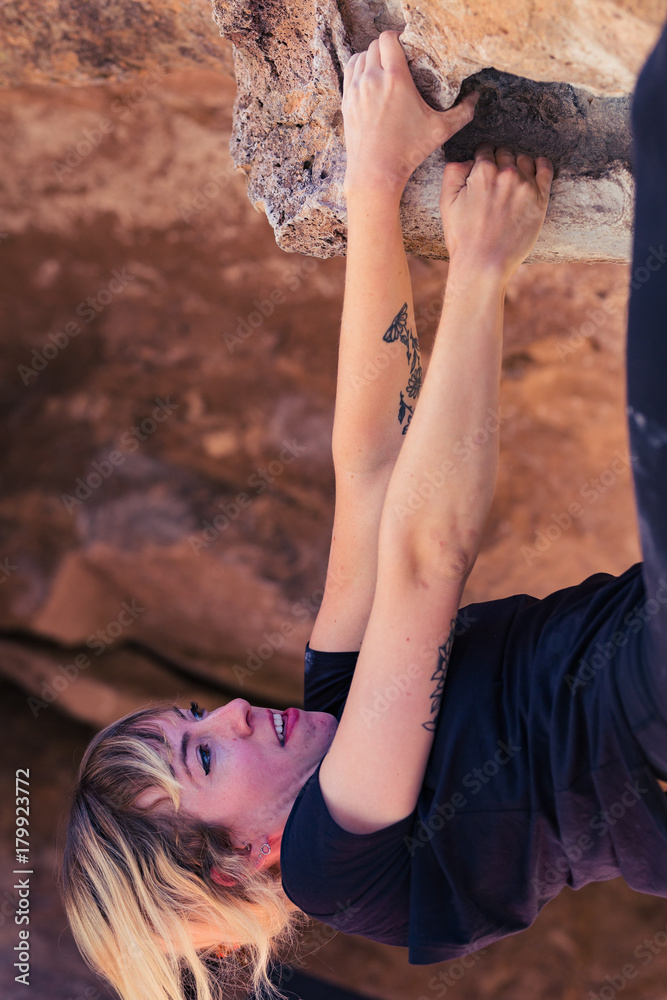 Naklejka premium Thin blonde caucasian woman with arm tattoos hangs upside down while she rock climbs on boulders in the desert of California