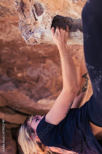 Thin blonde caucasian woman with arm tattoos hangs upside down while she rock climbs on boulders in the desert of California