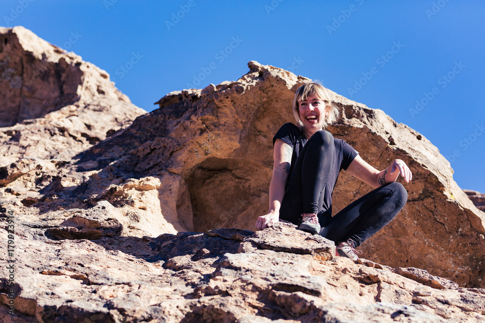 Thin blonde caucasian woman with arm tattoos sits triumphantly after rock climbing on boulders in the desert of California